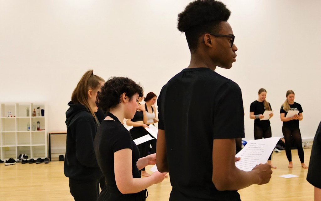 A circle of students all wearing black and they are holding pieces of white paper
