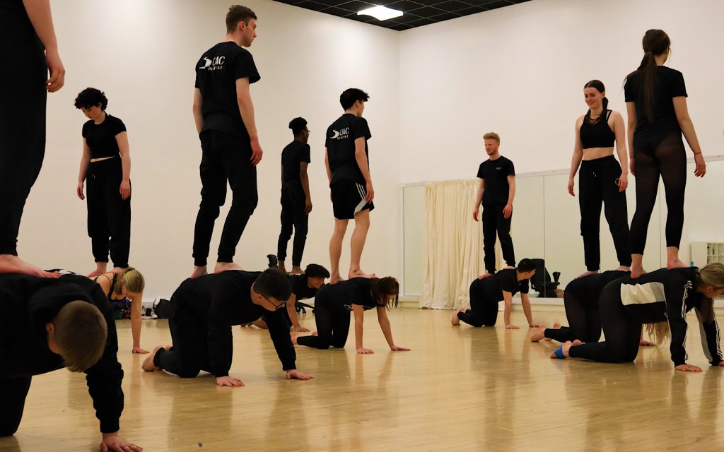Young dancers wearing black stand on top of each other’s backs in a workshop