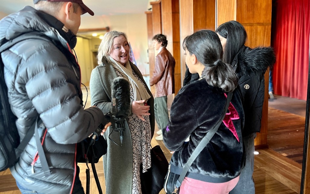 A woman with blonde hair is interviewed in the foyer of a theatre for a podcast