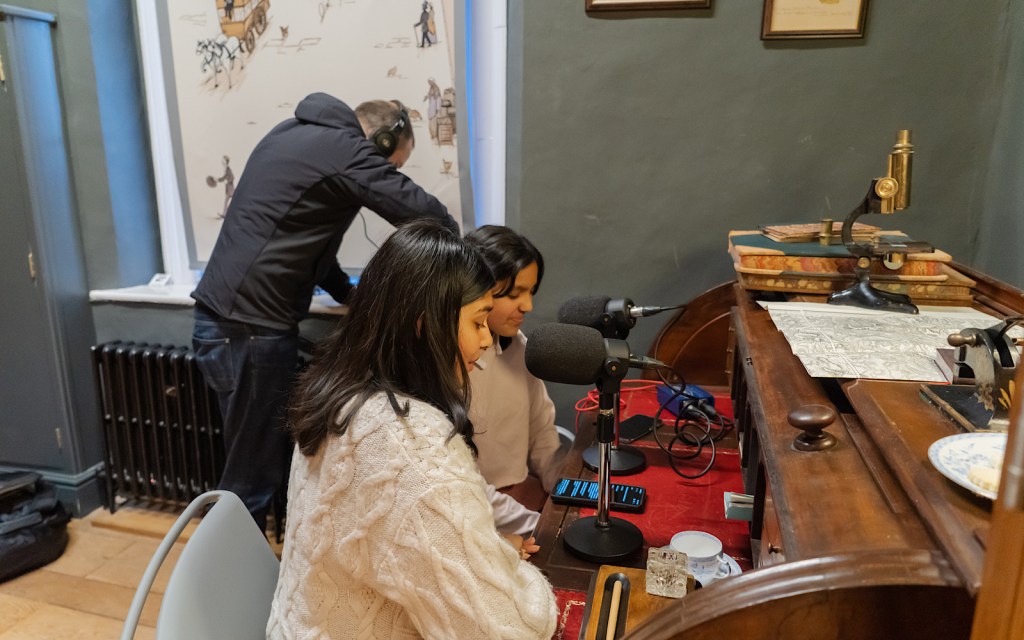 Two school students sit at a wooden desk talking into microphones whilst a podcast producer listens with headphones behind them