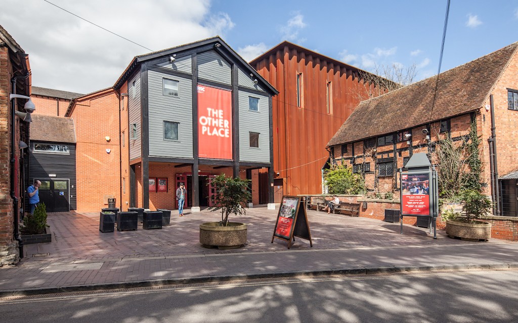 The outside of a theatre, made of wood and bricks and with a large metal warehouse attached to it
