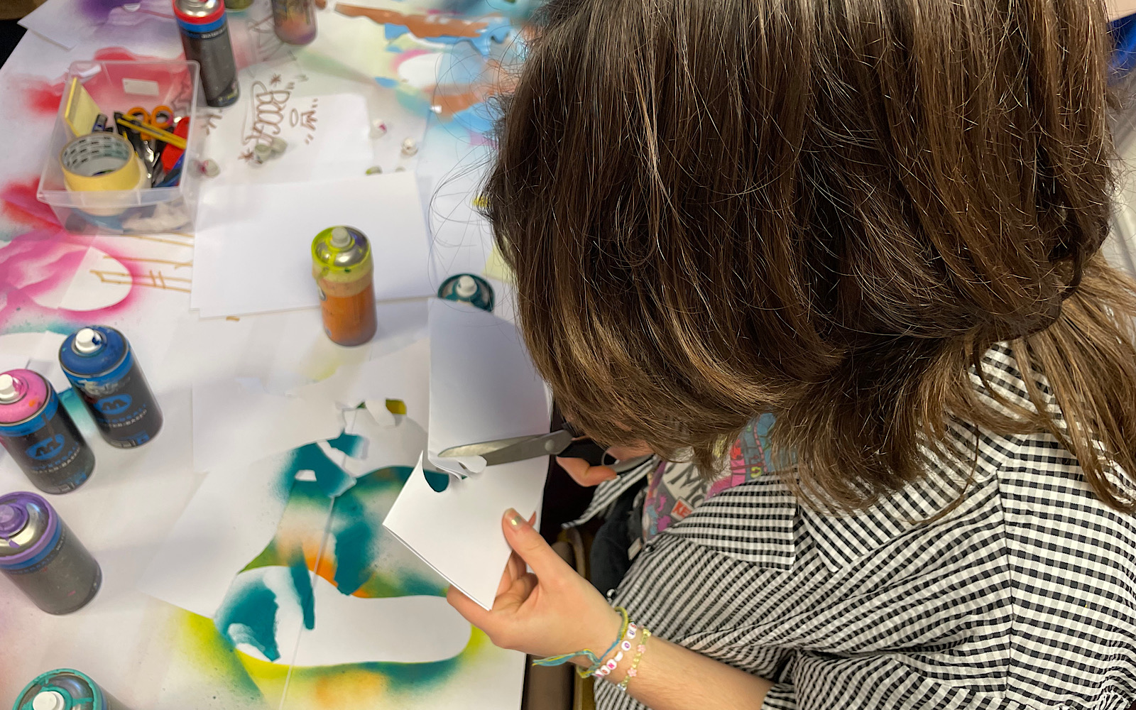 An artist with brown hair sits at a table and cuts out a stencil with scissors