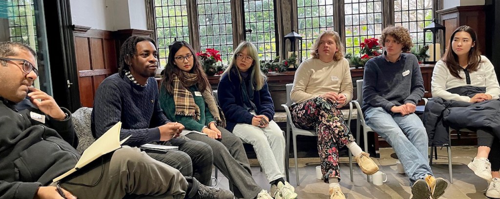 A group of seven students sit in a circle in a room talking to each other