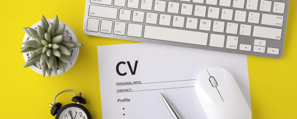 A CV sits on a yellow desk and is surrounded by a plant, a computer mouse, a clock and a keyboard