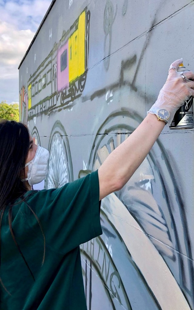 An artist in a green top is using a spray paint can against an outdoor wall