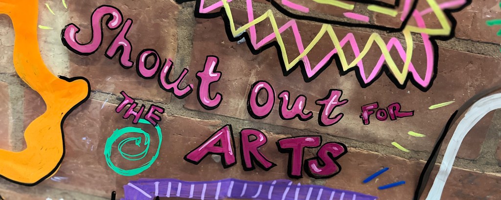 The words ‘shout out for the arts’ written in pink letters on a brick wall