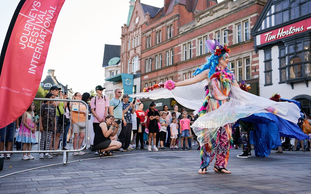 A dancer in a colourful costume is stood in a town centre with people from the town watching them