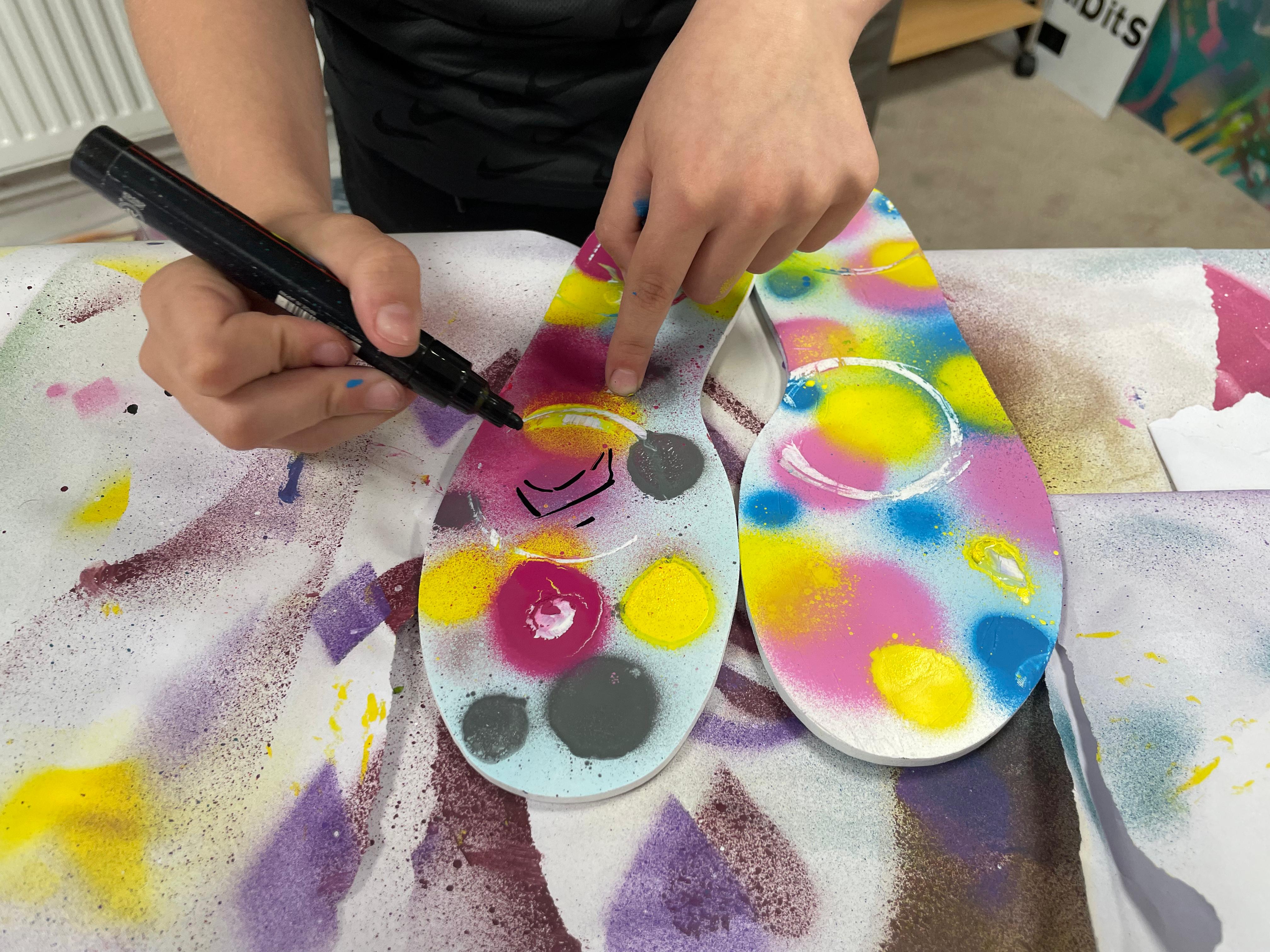 A child uses a marker to paint patterns on feet-shaped canvases on a table