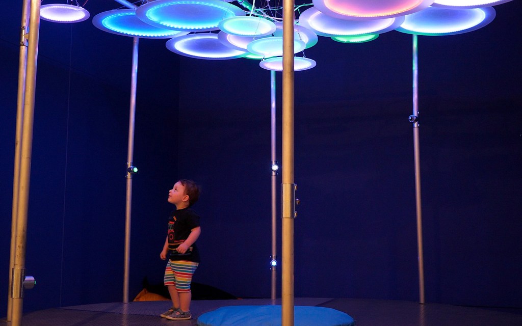 A child is looking at an illuminated art exhibition which is made of circular lights up above