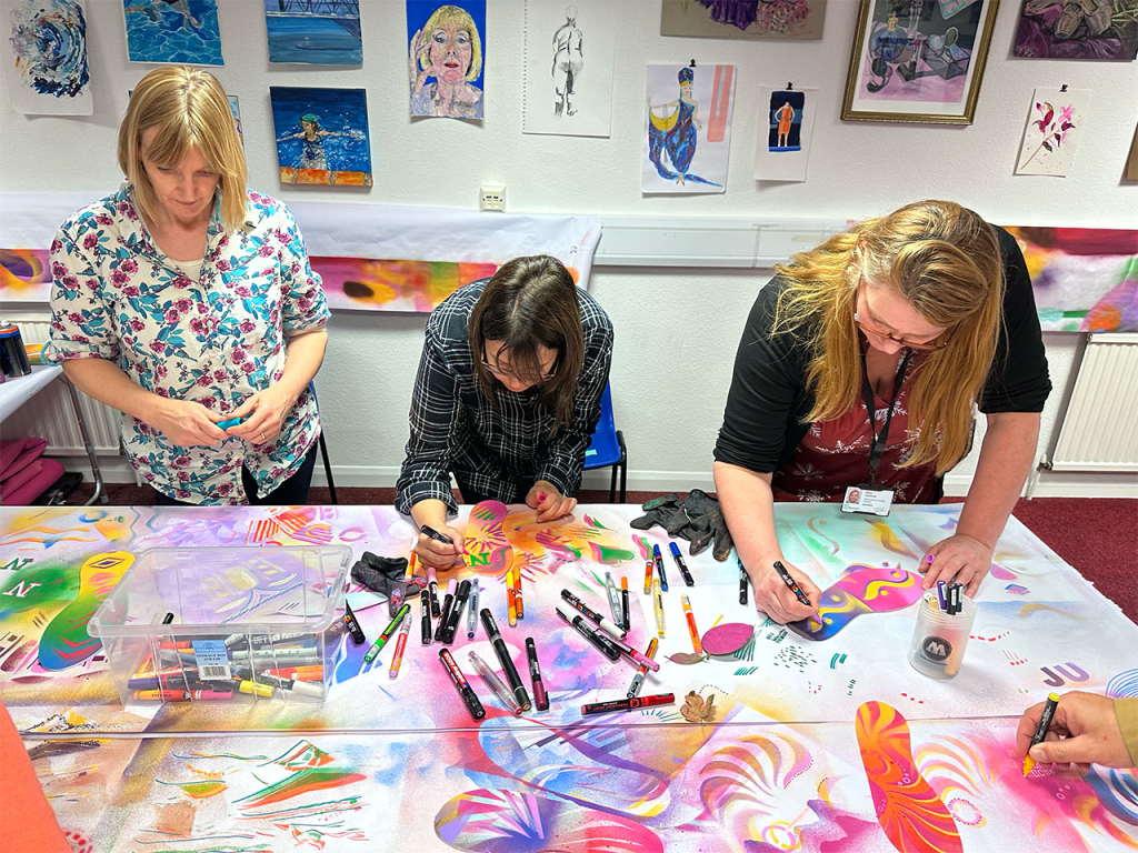 Three people take part in an art workshop, they lean over a table and use paints
