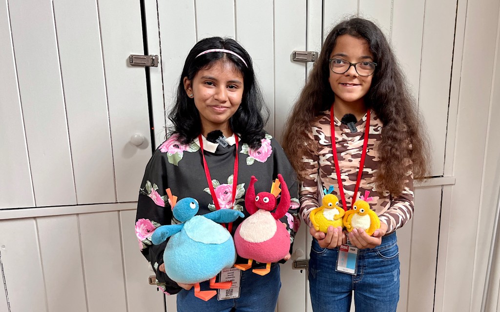 Two school students smile to the camera and they hold four chicken-like children’s toys in their hands, two of them yellow, one red and one blue
