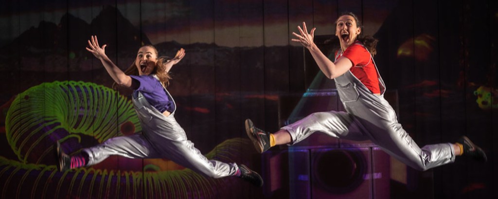Two dancers leap into the air as part of a performance and they are smiling
