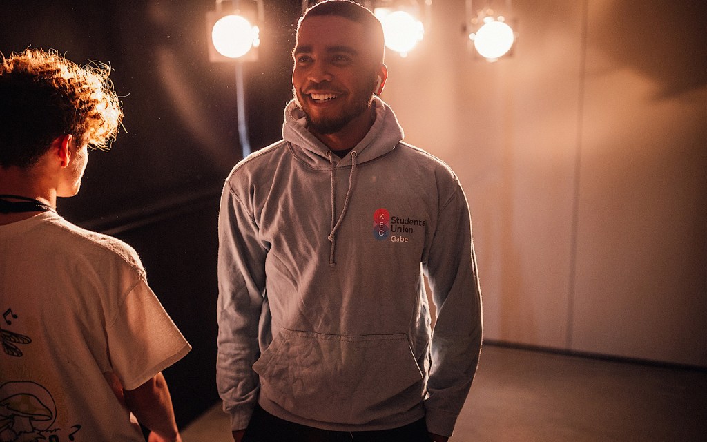 A student under spotlights wears a grey hoodie and they smile
