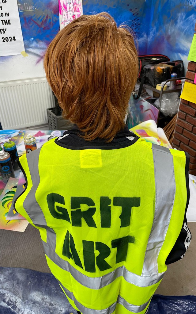 A child has a high vis jacket on and the words GRIT ART are spray painted on the back of it