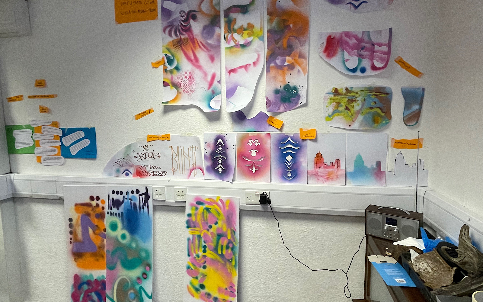 A wall has lots of paper stuck to it, on the paper is sketches, markings, stencils and patterns made with spray paint cans