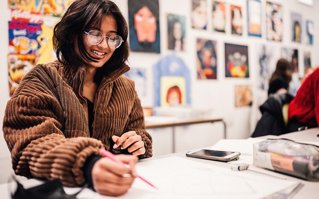 A student draws on a piece of paper, they smile and are wearing glasses