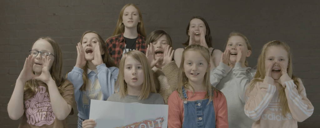 A group of school children hold up a sign which says shout out for the arts