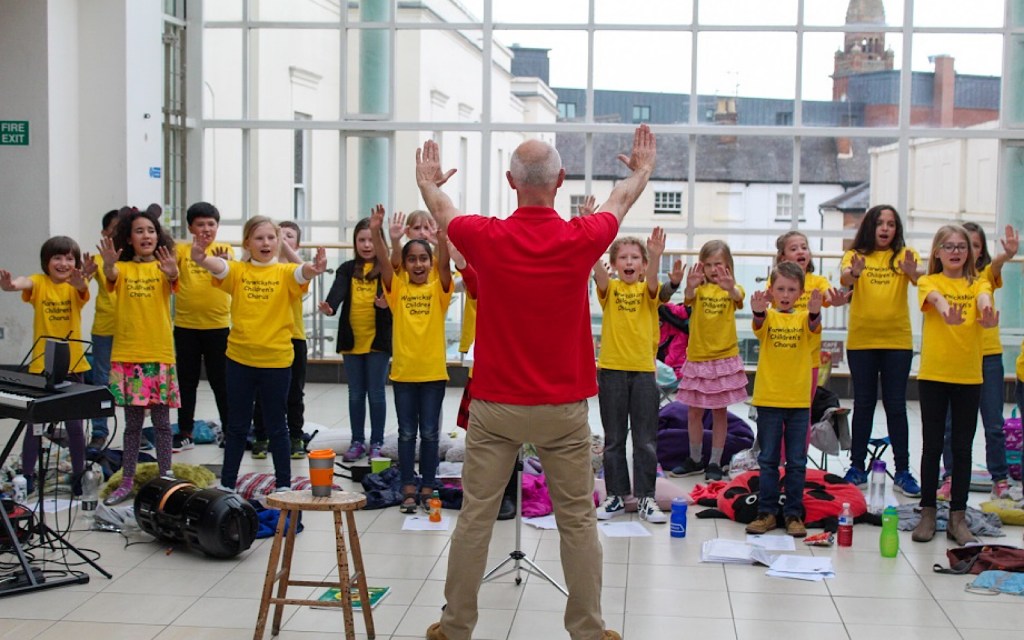 An adult instructs a group of children who are holding their arms in the air as part of a musical performance, they are all wearing yellow tshirts