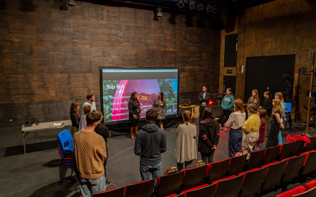 Young people stand in a studio theatre in a circle talking to one another