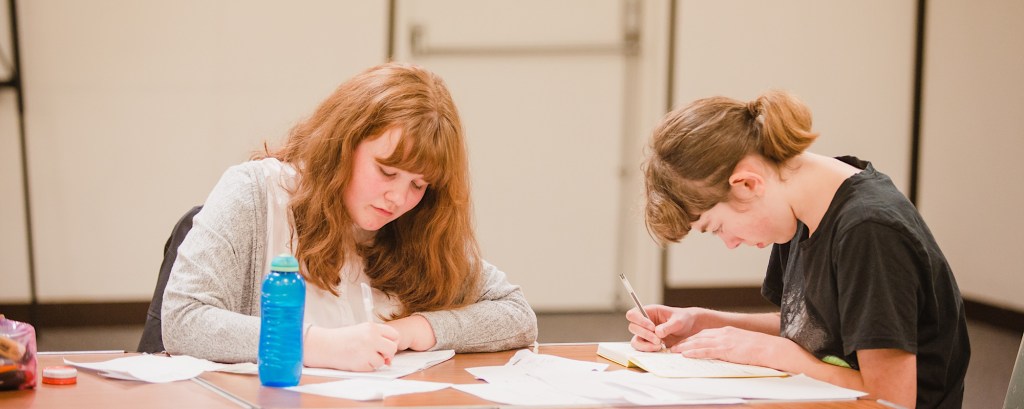 Two young people are at a wooden desk writing on pieces of paper