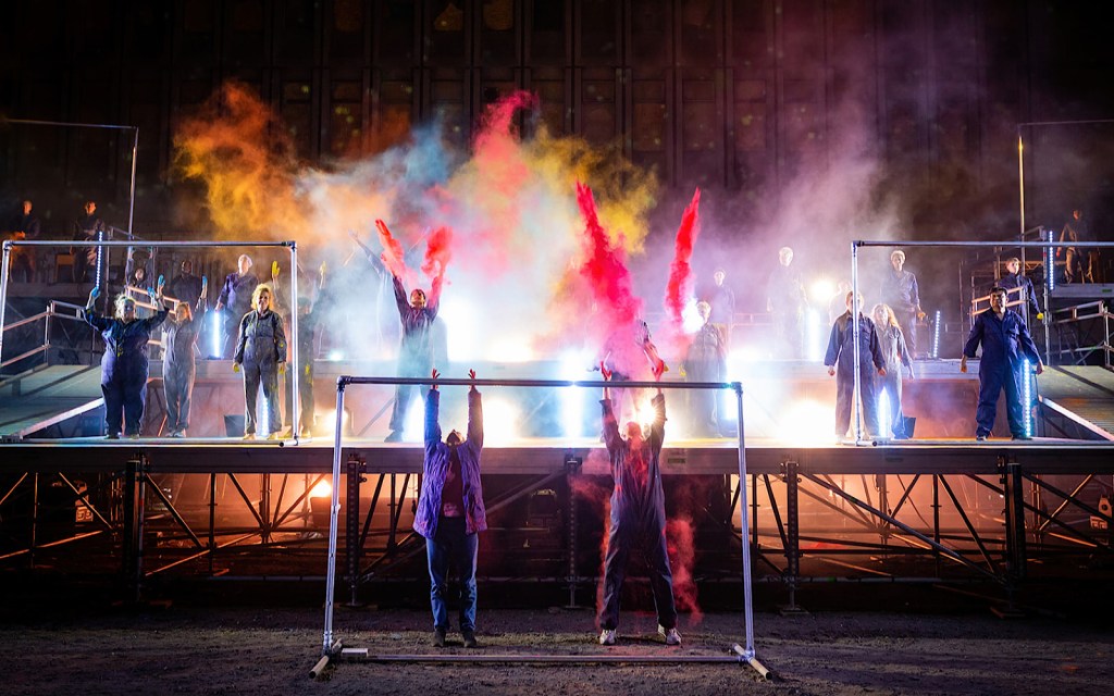 Performers on a stage in a town centre wear blue jumpsuits and fire coloured powder into the air