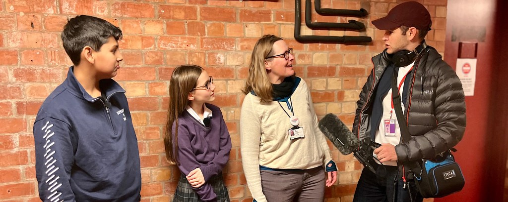 Two young people stand against a brick wall with two adults, the adults talk into a podcast microphone whilst the young people listen