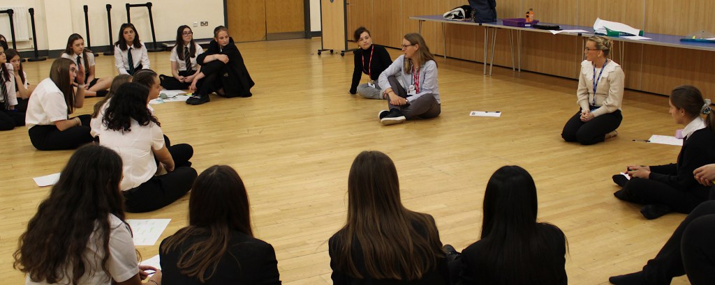 A group of students sit in a circle and listen to a practitioner who is telling them about Shout Out for the Arts