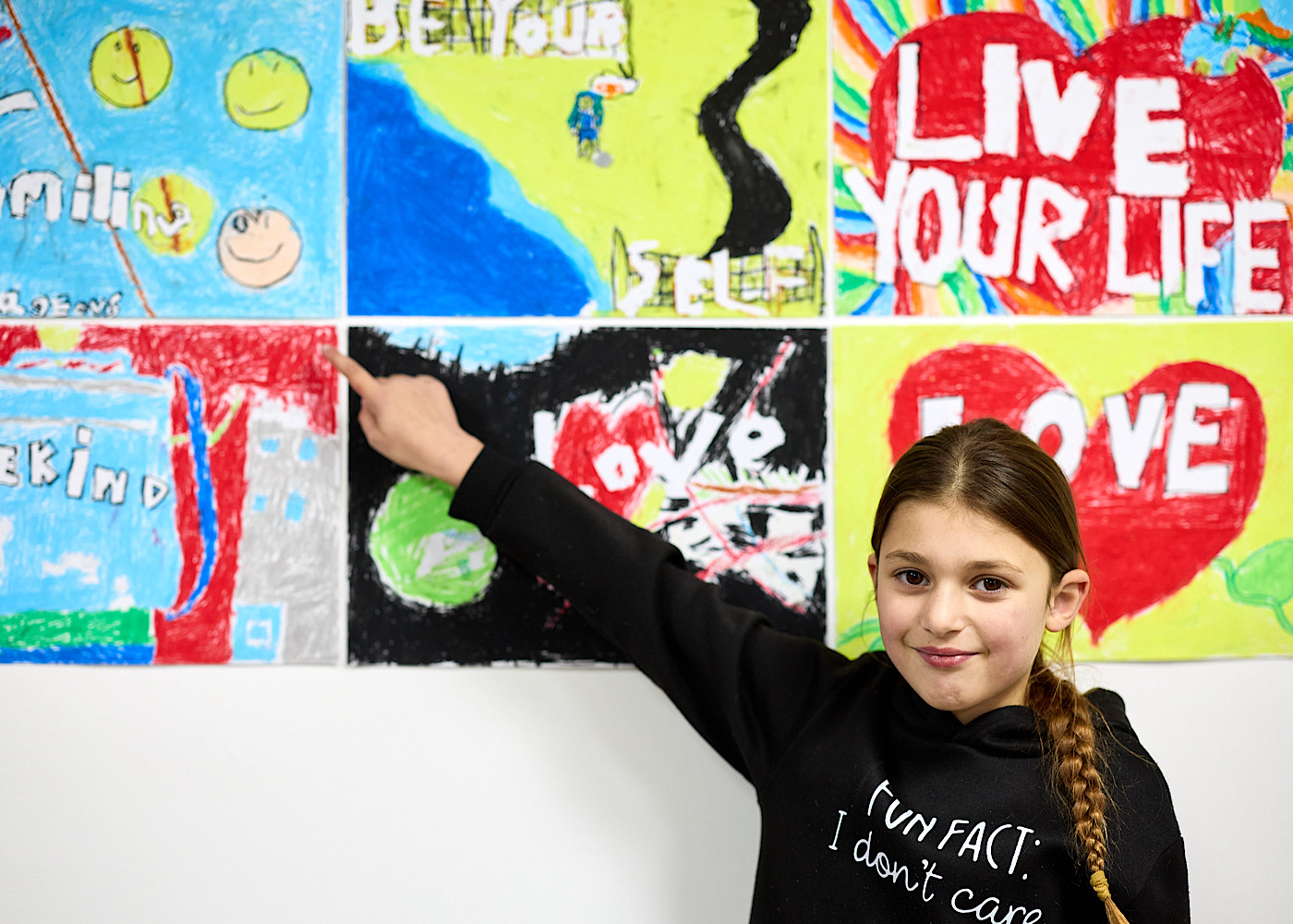 A young girl points to paintings on a wall which say motivational things like ‘live your life’ and ‘be yourself’