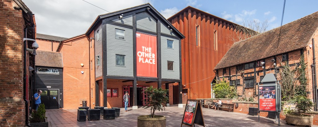 The outside of a theatre, made of wood and bricks and with a large metal warehouse attached to it