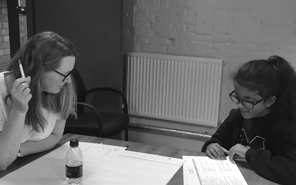 An adult and a child sit opposite each other on a table and they write notes