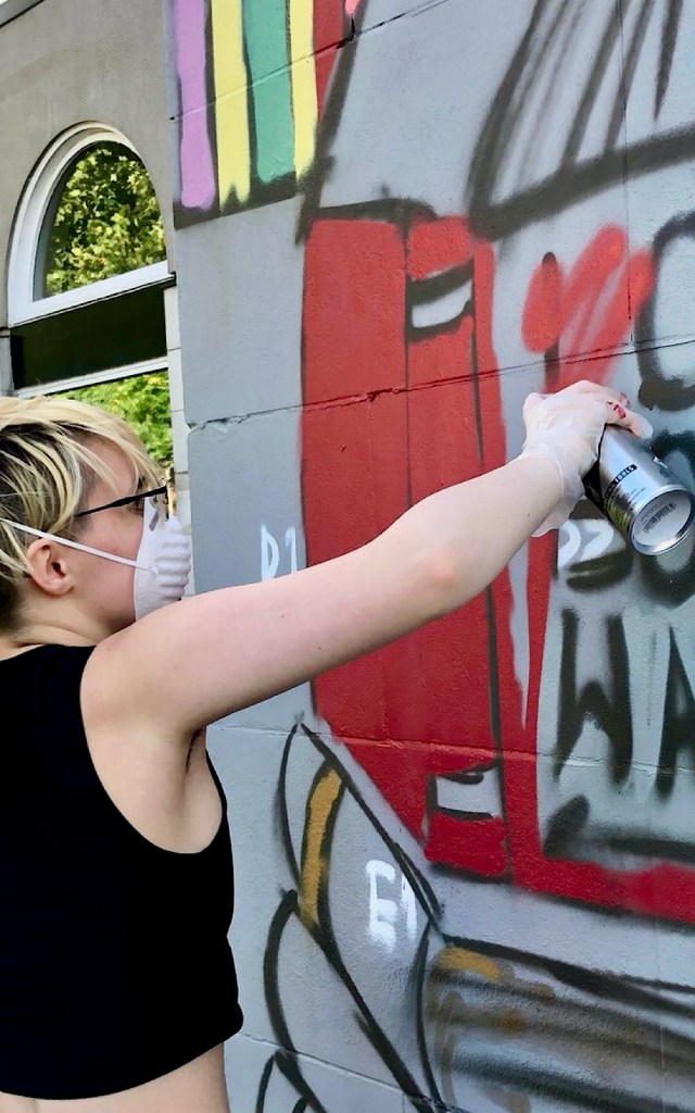 A young artist spray paints a book on a wall outside