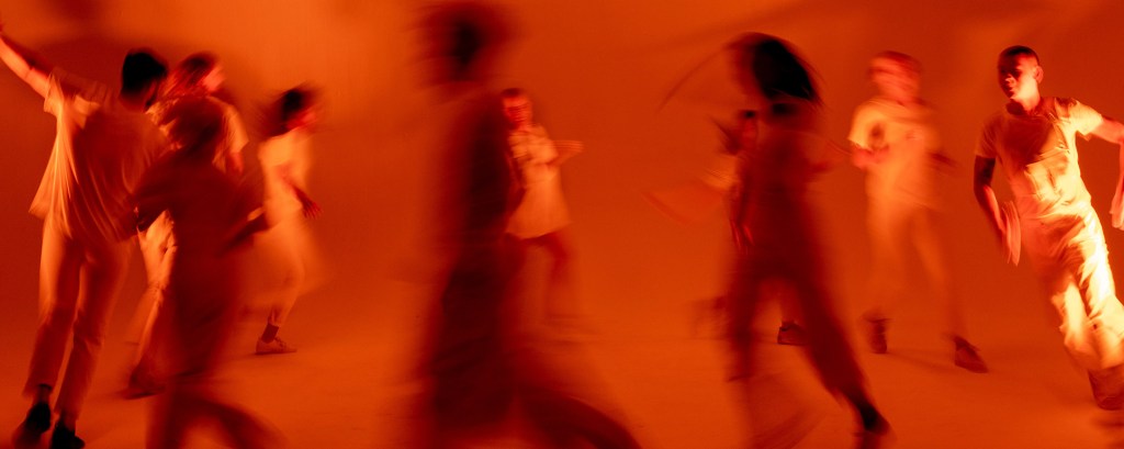 A group of people dance in a circle as part of a performance and the lighting is so red that the whole photo has a red wash over it