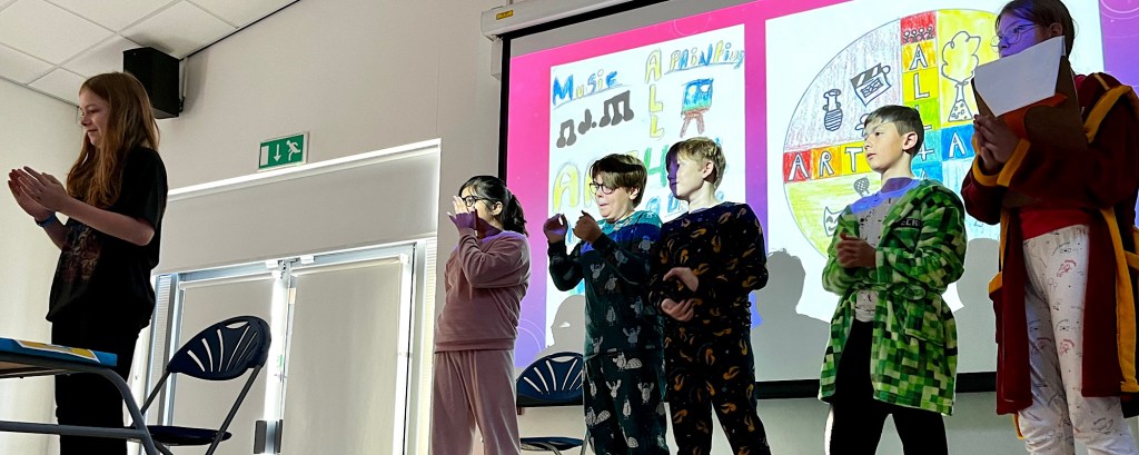 Six students stand on a stage for their school assembly and behind them is a presentation showing the words ‘arts for all’