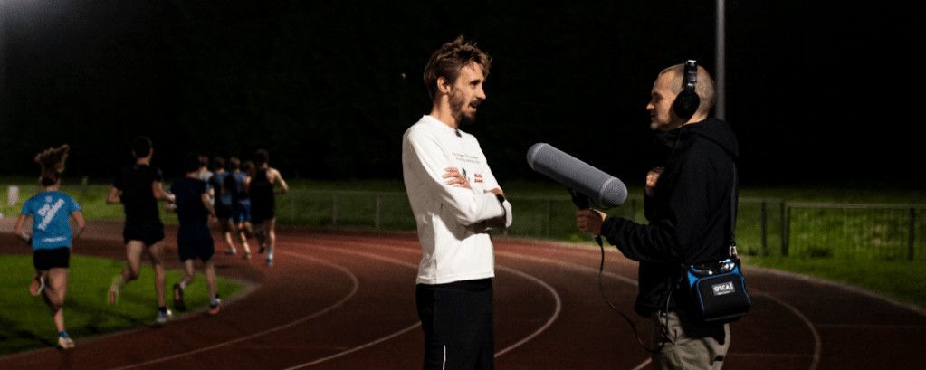 A podcast hosts interviews a person as they stand on a running track at night