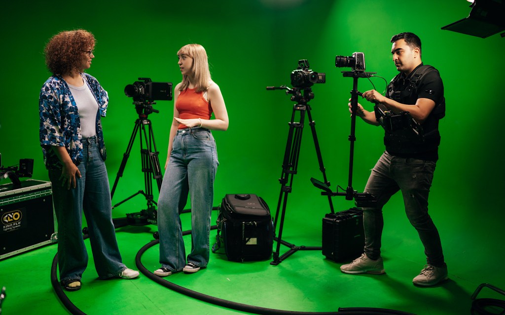 Three students stand in front of a green screen and film with cameras
