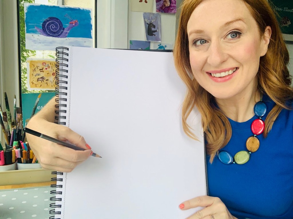An artist in a blue top smiles to the camera as she drawn on a pad with a pencil