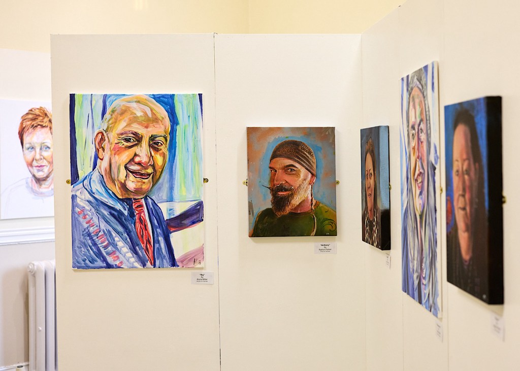 Six painted portraits of people’s faces are hung on a wall as part of an exhibition