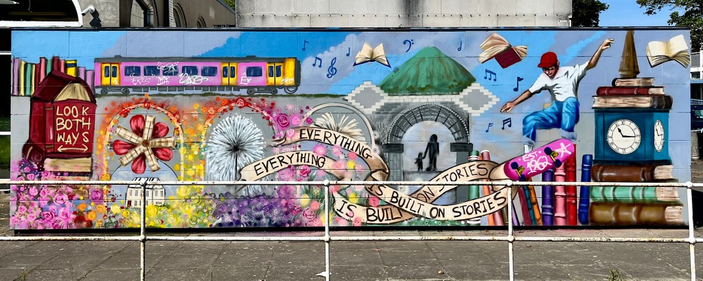 A mural on the side of a library in a town centre in Nuneaton, there’s a train, a skateboarder, a clock tower, books, flowers and the phrase ‘everything is built on stories’