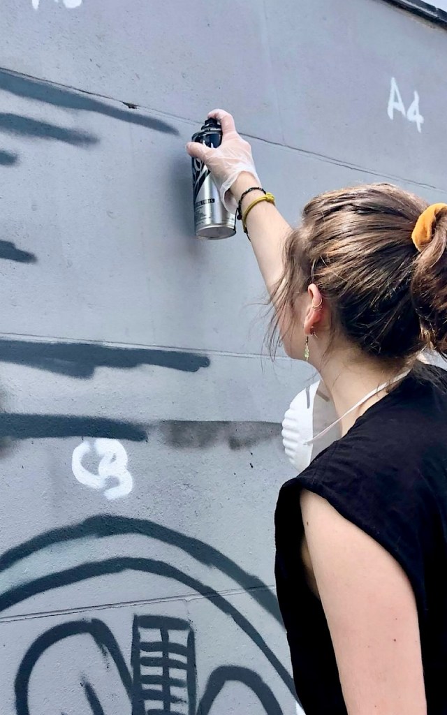 An artist holds a spray paint can against a grey wall