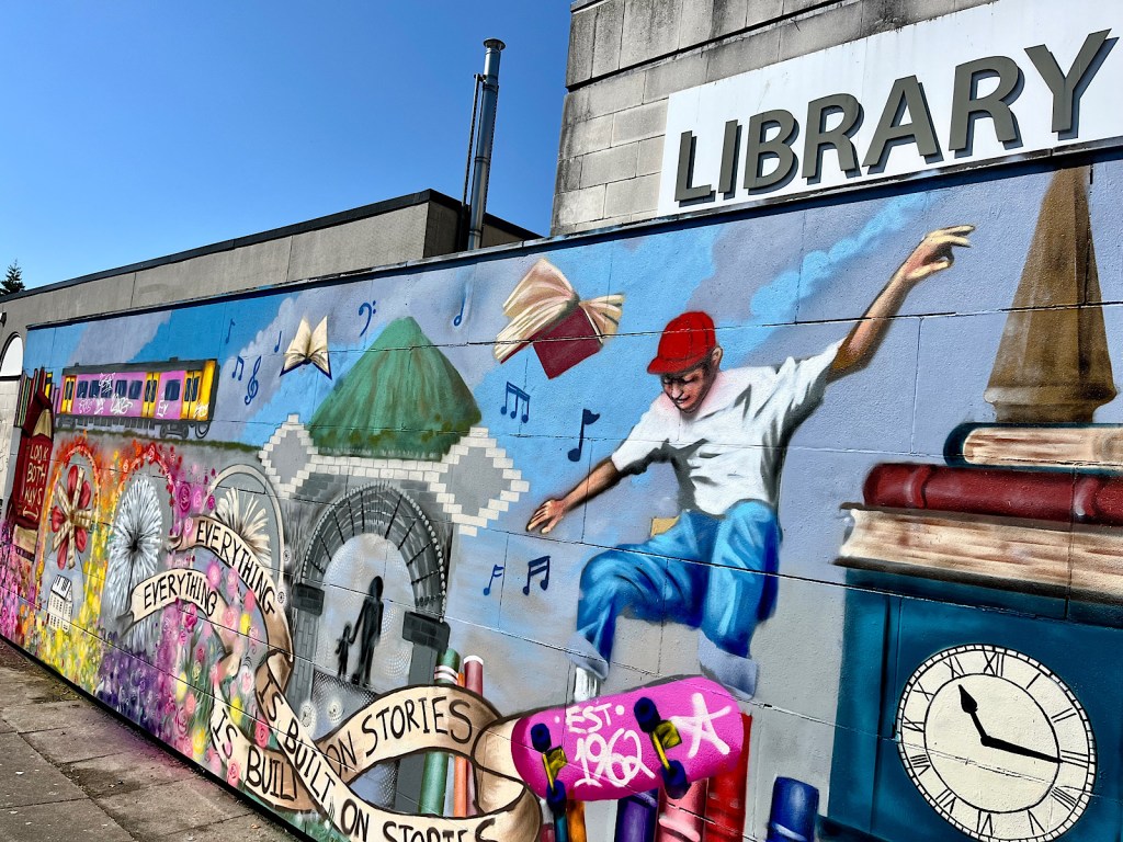 A mural on the side of a library in a town centre in Nuneaton, there’s a train, a skateboarder, a clock tower, books, flowers and the phrase ‘everything is built on stories’