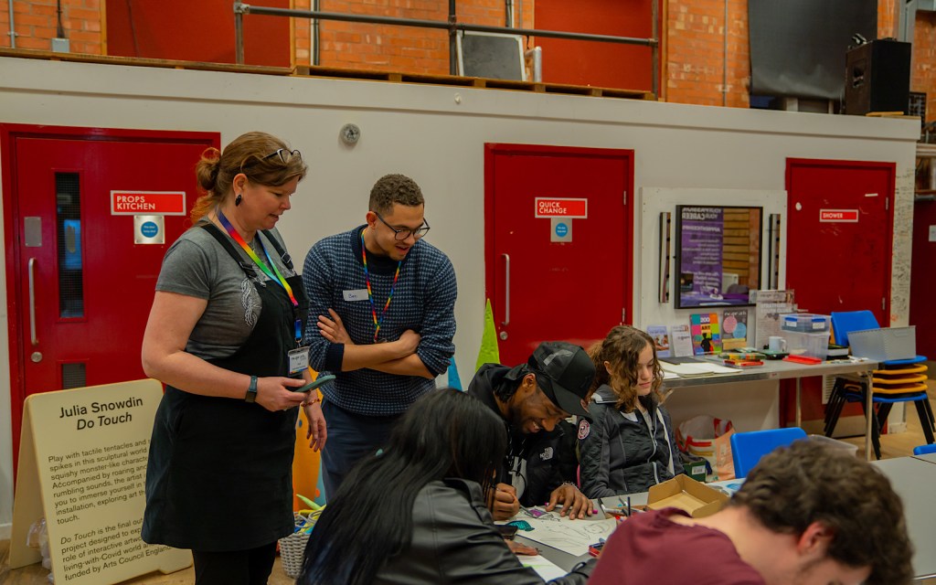 Two practitioners stand over students looking at the work they are creating in an art workshop