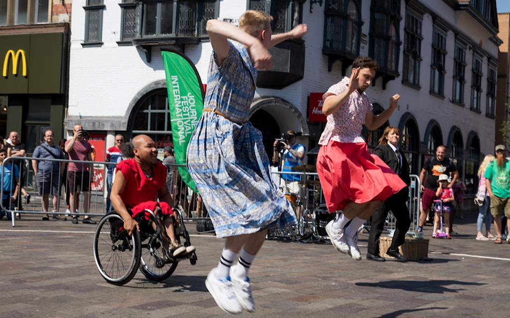 Three performers are in the middle of a town centre – two jump in the air and one uses a wheelchair