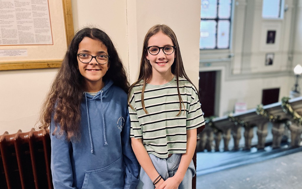 Two young people stand against a white wall and they both smile to camera