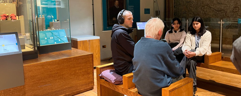 Two young students sit on wooden benches opposite a man holding podcast equipment and another person they are interviewing