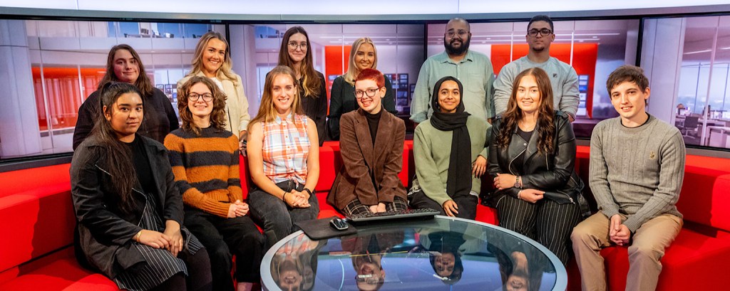 Two rows of apprentices look to the camera, at the front they sit on a red couch