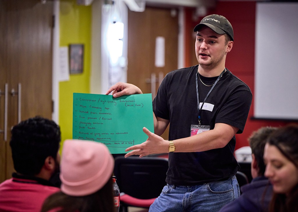 A person in a black t-shirt and cap shows a room of people a piece of paper with ideas written on it