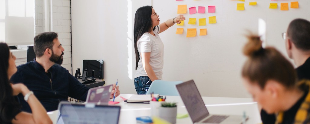 A group of young people sit at desks whilst an adult at the front sticks post-it notes onto a whiteboard