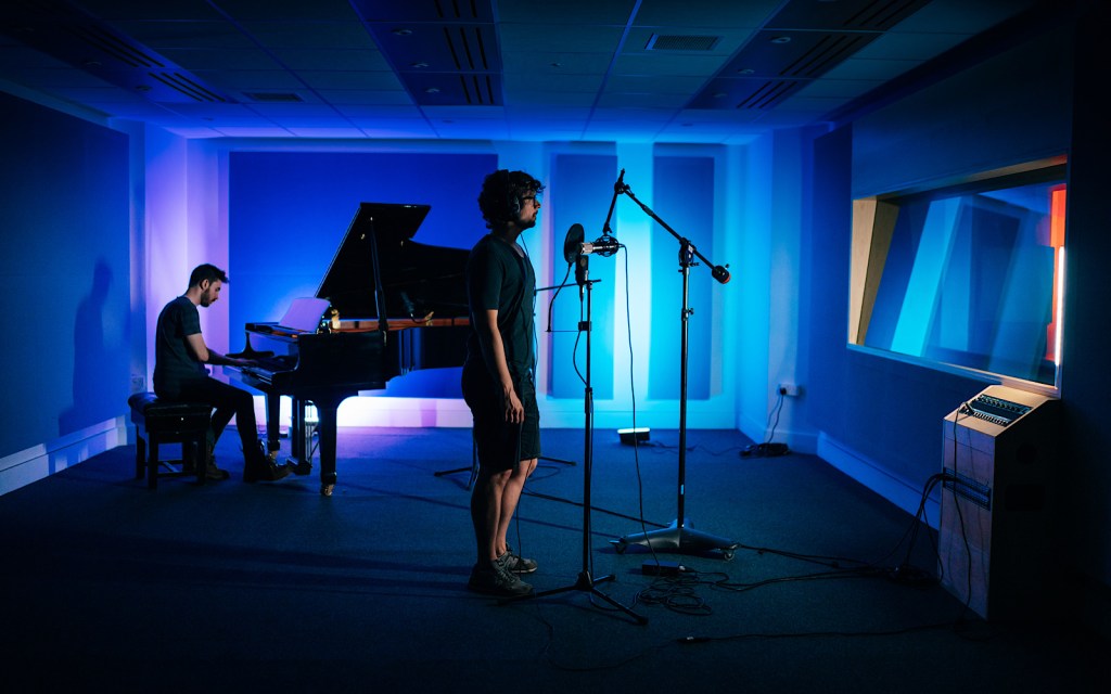 A student sings into a microphone and another one plays a piano in a recording studio