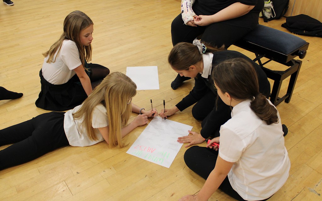 Students at a workshop with Shout Out for the Arts are writing on paper on a wooden floor