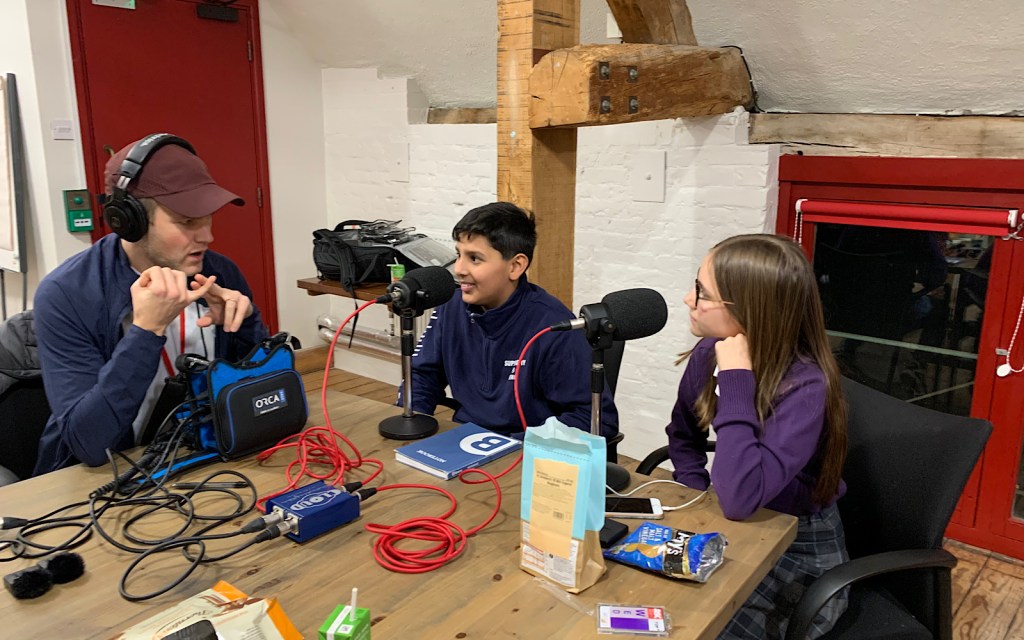Two school children sit at a wooden table and record a podcast, they are being guided by an adult podcast producer